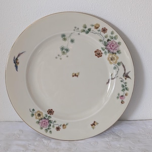 Op de afbeelding: Een crèmekleurige porseleinen bord met een gouden rand. Het bord heeft bloemen- en vogelontwerpen in roze, geel, bruin en blauw. Er zijn ook vlinders afgebeeld. Het bord is een decoratief stuk om te serveren of tentoon te stellen.