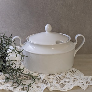 Può includere: Una zuppiera in ceramica bianca con coperchio e due manici. La zuppiera è appoggiata su un centrino di pizzo bianco. Il coperchio ha un piccolo pomello e una fessura per il cucchiaio. Lo sfondo è grigio neutro.