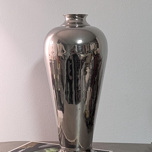 Könnte beinhalten: Eine hohe, polierte silberfarbene Metallvase mit einer glatten, reflektierenden Oberfläche. Die Vase hat einen sich verjüngenden Körper, der sich zur Schulter hin verbreitert, einen schmalen Hals und einen ausgestellten Rand. Sie steht auf einem runden Fuß.