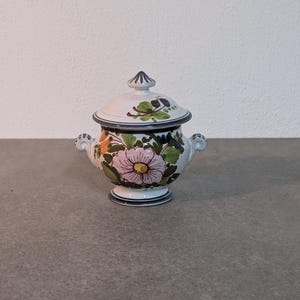 Op de afbeelding: Een klein wit suikerpotje van keramiek met een deksel, versierd met een bloemenmotief in de kleuren roze, geel, groen en bruin. Het potje heeft twee handvatten en een ronde basis.