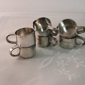 Könnte beinhalten: Mehrere kleine, silberfarbene Metallbecher mit Henkeln sind paarweise gestapelt. Die Becher haben ein schlichtes, klassisches Design. Einige Becher haben Text auf der Seite. Die Becher sind auf einer weißen Oberfläche angeordnet.