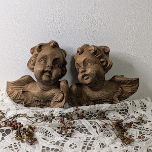 Puede incluir: Dos esculturas de ángeles de madera con tallas detalladas de rostros y alas. Los ángeles son de color marrón cálido y están colocados uno al lado del otro. Descansan sobre un mantel de encaje blanco, con una ramita de bayas delante.