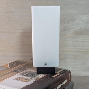 Könnte beinhalten: Eine weiße Tischlampe mit schwarzem Fuß. Die Lampe steht auf einem Stapel Bücher.