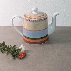 Könnte beinhalten: Eine Keramik-Teekanne mit einem mehrfarbigen horizontalen Streifenmuster in Beige, Hellblau, Dunkelgrün und Orange. Der Deckel hat einen kleinen Knaufgriff und eine florale Bordüre. Sie verfügt über einen geschwungenen Henkel und einen Ausguss.