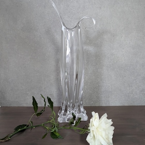 Könnte beinhalten: Eine klare Glasvase mit einem einzigartigen, geschwungenen Design. Die Vase hat eine hohe, schlanke Form mit einer breiten Öffnung oben. Die Vase steht auf einer Holzoberfläche mit einer einzelnen weißen Rose und einem grünen Stiel.