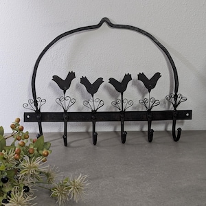 Puede incluir: Un perchero de pared de metal negro con cinco ganchos. El perchero presenta cuatro siluetas de pájaros y adornos decorativos. La parte superior del perchero tiene un diseño curvo. El perchero está montado sobre una superficie gris.