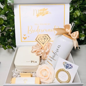 Puede incluir: Una caja de regalo blanca con detalles dorados, que contiene un vaso, una caja de joyas, una scrunchie y otros artículos. La tapa de la caja dice "Madison, Will You Be My Bridesmaid?" en letras doradas. También se incluye una pegatina de diamante dorado y una rosa artificial.