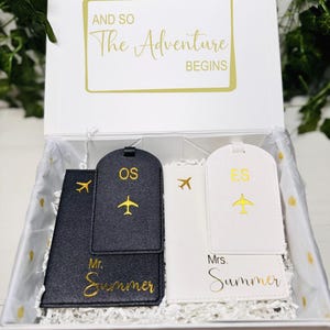 Puede incluir: Una caja de regalo blanca con el texto dorado "AND SO The Adventure BEGINS". En el interior, etiquetas de equipaje y portapasaportes negros y blancos con diseños de aviones dorados. Las etiquetas están personalizadas con iniciales y el apellido "Summer".