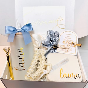 Könnte beinhalten: Eine weiße Geschenkbox gefüllt mit Artikeln für eine Brautjungfer. Die Box enthält einen Becher mit Strohhalm, ein Haargummi, eine Haarklammer, eine Schmuckbox und eine Perlenhaarklammer. Der Becher und die Schmuckbox sind mit dem Namen "Laura" personalisiert. Die Karte sagt "Willst du meine Brautjungfer sein?" und "Zu haben und zu halten, deine Haare sehen toll aus".