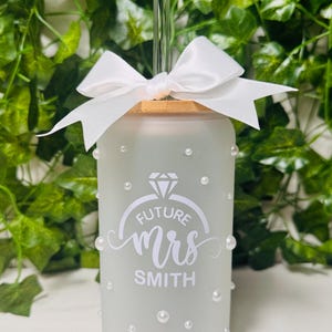 May include: A frosted glass tumbler with a glass straw and a white bow. The tumbler is decorated with white pearls and the words "FUTURE MRS SMITH" inside a ring design. The tumbler is a wedding-themed product.
