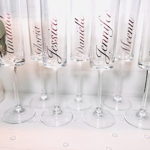 Op de afbeelding: Transparante champagneglazen met namen in een sierlijk lettertype. De namen Amanda, Gloria, Jessica, Danielle, Jennifer, Meenu en Sherry zijn zichtbaar. De glazen staan op een witte ondergrond.