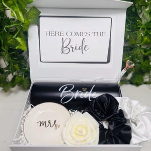 Puede incluir: Una caja de regalo blanca con un tema en blanco y negro, que contiene un vaso negro con "Bride" en escritura blanca, un plato pequeño con "mrs" en escritura dorada y coleteros blancos y negros. La caja también incluye un bolígrafo con una parte superior en forma de diamante y una tarjeta blanca que dice "Here Comes The Bride".