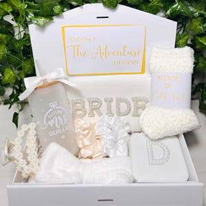 May include: A white gift box containing bridal accessories. It features a frosted glass with "Future Mrs. Guillen", a pearl hair clip, scrunchies, socks, and a box with the letter "D". The box also has the text "And So The Adventure Begins".