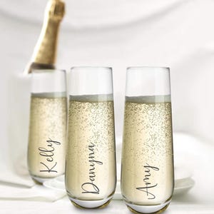 Op de afbeelding: Drie doorzichtige champagneglazen zonder steel, gevuld met mousserende vloeistof. Elk glas is voorzien van een naam in zwarte cursieve letters: Kelly, Danyna en Amy. De glazen hebben een cilindrische vorm met een licht toelopende onderkant.