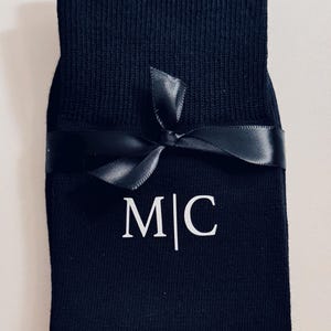Puede incluir: Un par de calcetines negros con una cinta negra atada alrededor. Los calcetines tienen las iniciales "M|C" bordadas en blanco.