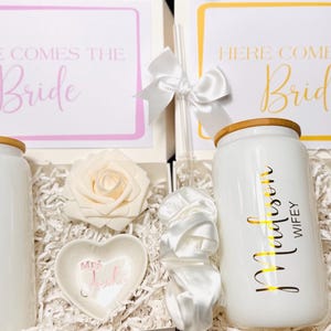 Puede incluir: Dos cajas de regalo con artículos temáticos de boda. Cada caja contiene un vaso blanco con tapa de madera, un plato en forma de corazón y una rosa decorativa. Una caja tiene un letrero con marco rosa que dice "Here Comes the Bride".