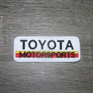May include: White rectangular embroidered patch with black text that reads "TOYOTA MOTORSPORTS". The text is outlined in yellow and red.