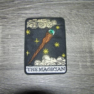 May include: A black and gold embroidered patch with a wand and stars. The text "THE MAGICIAN" is at the bottom of the patch.