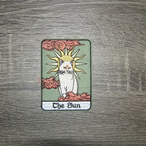May include: A green and pink tarot card with a white cat in the center. The cat has a third eye and is surrounded by a yellow sun. The card reads "The Sun".