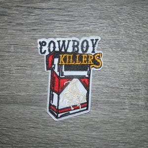 May include: An embroidered patch featuring a rectangular cigarette pack design with red, white, and black colours. The text COWBOY KILLERS is stitched in black and gold above the pack, which displays a white horse and rider graphic in the centre.
