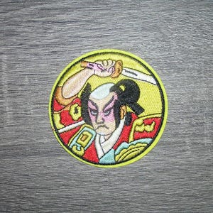May include: Embroidered patch featuring a stylized illustration of a samurai warrior with a sword. The patch is circular, with a yellow background and a black border. The warrior is depicted in traditional attire, with a stern expression.