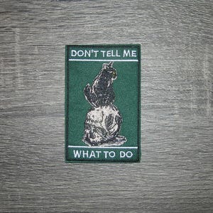 May include: Embroidered patch with a black cat illustration on a green background. The patch has the text "DON'T TELL ME" at the top and "WHAT TO DO" at the bottom in white lettering. The cat is sitting on a ball.