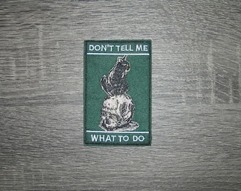 May include: Embroidered patch with a black cat illustration on a green background. The patch has the text "DON'T TELL ME" at the top and "WHAT TO DO" at the bottom in white lettering. The cat is sitting on a ball.