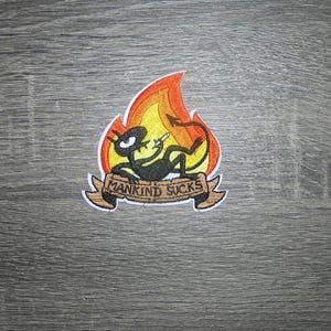 May include: Embroidered patch featuring a cartoon devil character in flames, holding a cigarette. The patch has a white border and a banner that reads "MANKIND SUCKS" in brown lettering. The design is on a gray wooden surface.