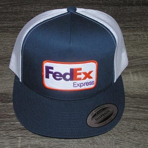 May include: Navy blue and white trucker hat with the FedEx Express logo on the front. The hat features a mesh back and a curved brim. The logo is a rectangular patch with the FedEx logo in purple and orange.