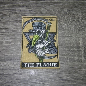 May include: Embroidered patch featuring a skeletal figure in a plague doctor mask, holding a scythe. The design includes the text "THE PLAGUE" and the Roman numeral "XIII" on a tan background.