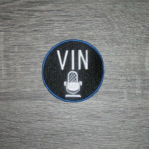 May include: A circular embroidered patch with a black background and a thin blue border. The patch features the white text VIN positioned above a white graphic illustration of a vintage-style microphone centered on the black fabric surface.