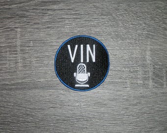 May include: A circular embroidered patch with a black background and a thin blue border. The patch features the white text VIN positioned above a white graphic illustration of a vintage-style microphone centered on the black fabric surface.