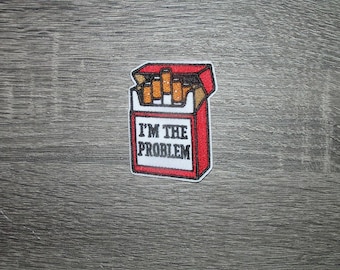 May include: Embroidered patch featuring a red and white cigarette pack with the text "I'M THE PROBLEM" in black. The pack is open, revealing several cigarettes. The patch has a white border and is set against a gray wood grain background.