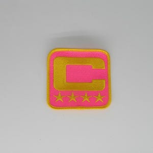 Peut inclure: Un écusson brodé carré avec un fond rose vif et une bordure jaune. Le centre présente une grande lettre C jaune, et sous la lettre se trouvent quatre petites étoiles brodées jaunes disposées sur une ligne horizontale.