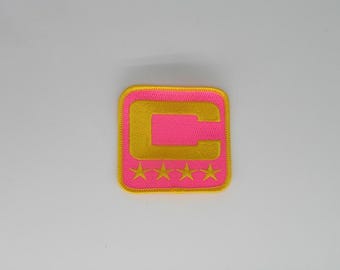 May include: A square embroidered patch featuring a bright pink background with a yellow border. The center displays a large yellow letter C, and below the letter are four small yellow embroidered stars arranged in a horizontal row.