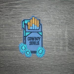 Peut inclure: Écusson brodé représentant un paquet de cigarettes bleu clair avec le texte "COWBOY SOULS" en bleu foncé. Les cigarettes sont de couleur beige et bleue. Des motifs floraux turquoise sont en bas. L'écusson est sur un fond en bois gris.