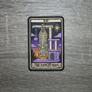 May include: A rectangular embroidered patch featuring a tarot card design titled The Hanged Man. A tabby cat hangs upside down from a cat tree structure against a purple and orange sunset background with a yellow star and the Roman numeral XII.