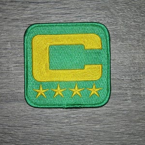 Op de afbeelding: Geborduurde patch met een groene vierkante achtergrond en een gele letter "C" in het midden. Onder de letter staan vier gele sterren. De patch is waarschijnlijk voor kleding of accessoires.