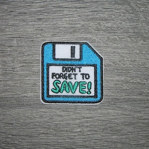 Może przedstawiać: Niebieska, biała i czarna haftowana naszywka w kształcie dyskietki. Na naszywce widnieje napis "Didn't Forget to SAVE!" w zabawnym kroju pisma. Naszywka ma białą obwódkę i jest umieszczona na szarym tle.