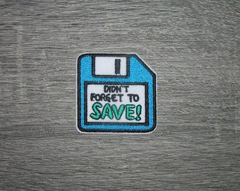May include: A blue, white, and black embroidered patch in the shape of a floppy disk. The patch reads "Didn't Forget to SAVE!" in a playful font. The patch has a white border and is set against a gray background.
