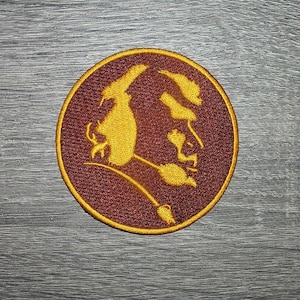 May include: A circular embroidered patch featuring a deep maroon background with a gold-colored border and a stylized gold graphic of a profile silhouette of a person with a feathered headdress, rendered in detailed thread work.