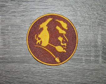 May include: A circular embroidered patch featuring a deep maroon background with a gold-colored border and a stylized gold graphic of a profile silhouette of a person with a feathered headdress, rendered in detailed thread work.