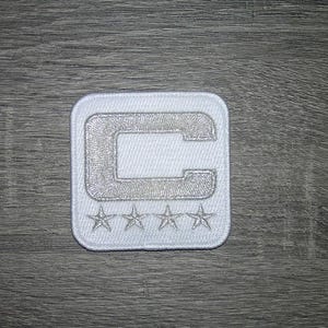 May include: A square white embroidered patch featuring a large capital letter C in the center. Below the letter C, there is a horizontal row of four small, five-pointed stars. The entire design is stitched in white thread on a white fabric base.