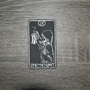 May include: Black and white embroidered patch of the tarot card The Hermit. The Hermit is depicted as a hooded figure holding a lantern. The card number IX is visible in the top right corner.