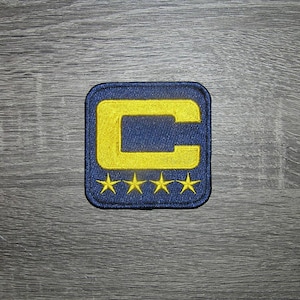 May include: Embroidered patch featuring a navy blue square with a yellow "C" and four yellow stars. The patch is likely for apparel or accessories, adding a decorative element. The design is simple and bold.