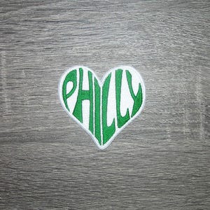 May include: A heart-shaped patch with the word "PHILLY" embroidered in green on a white background. The patch is placed on a gray wooden surface. The patch is likely a souvenir or a decorative item.