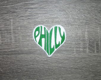 May include: A heart-shaped patch with the word "PHILLY" embroidered in green on a white background. The patch is placed on a gray wooden surface. The patch is likely a souvenir or a decorative item.