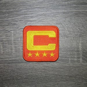 Op de afbeelding: Geborduurde patch met een vierkante vorm en een rode achtergrond. Een gele letter "C" staat in het midden, omrand met rood. Vier gele sterren staan onderaan. De patch ligt op een grijs houten oppervlak.
