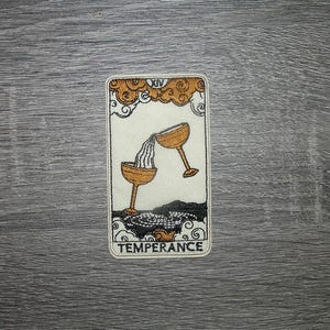 May include: Embroidered patch featuring the Temperance tarot card. The design shows two golden chalices with water flowing between them, set against a cream background. The card is framed with black and gold accents, and the word "TEMPERANCE" is at the bottom.