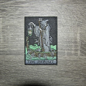Op de afbeelding: Een zwart en groen geborduurde patch met een gekapte figuur die een lantaarn vasthoudt en in een boot staat. De tekst "The Hermit" staat onderaan de patch.